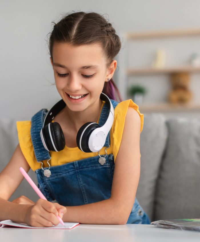 Lifestyle Concept. Smiling school girl with colorful dyed hair sitting at desk, writing essay in copybook. Pupil with headset studying and doing homework or taking notes in her diary, making checklist